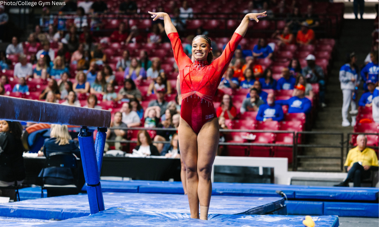 Gabrille Dildy, a gymnast from Rutgers University, wearing a red leotard stands on a blue mat in a large arena, with her arms raised in a "V" shape and a wide smile. She is positioned in front of a balance beam, with a crowd of spectators in red and blue seating visible in the blurred background.