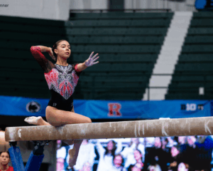 A gymnast poses on the balance beam in a sitting position. One hand is outstretched and the other is behind her head. She wears a red and black leotard.