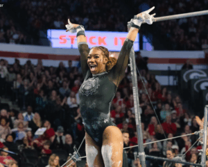 Here is the alt text for the image: **Alt Text:** A gymnast, Ja'Free Scott of the University of Georgia, wearing a black leotard, raises her chalk-covered arms in celebration after finishing a routine on the uneven bars. She is smiling excitedly with her mouth open, standing in front of the equipment and a large crowd in an arena.
