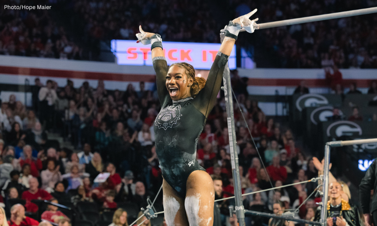 Here is the alt text for the image: **Alt Text:** A gymnast, Ja'Free Scott of the University of Georgia, wearing a black leotard, raises her chalk-covered arms in celebration after finishing a routine on the uneven bars. She is smiling excitedly with her mouth open, standing in front of the equipment and a large crowd in an arena.