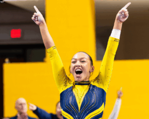 A gymnast salutes, pointing up into the air and she smiles wide over a yellow background. She wears a blue and yellow long-sleeved leotard and her hair is pulled back.