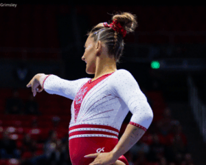 A gymnast stands against a black background looking away from the camera. One hand is on her hip and the other pointing into the distance. Her hair is braided into an updo and she wears a white and red leotard.