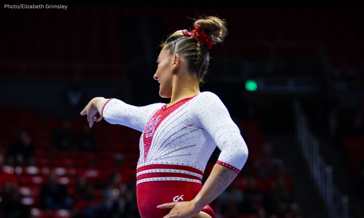 A gymnast stands against a black background looking away from the camera. One hand is on her hip and the other pointing into the distance. Her hair is braided into an updo and she wears a white and red leotard.