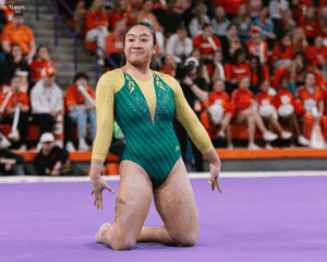 Michelle Ngo, a female gymnast from William & Mary, finishes a floor routine on a purple mat. She is kneeling with her chest up and arms held slightly back, wearing a green and gold long-sleeved leotard with sparkly accents. She has a focused expression and blue glitter on her cheek. In the background, a crowd of spectators in orange clothing sits in the bleachers of an arena.