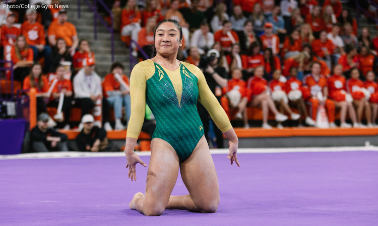 Michelle Ngo, a female gymnast from William & Mary, finishes a floor routine on a purple mat. She is kneeling with her chest up and arms held slightly back, wearing a green and gold long-sleeved leotard with sparkly accents. She has a focused expression and blue glitter on her cheek. In the background, a crowd of spectators in orange clothing sits in the bleachers of an arena.