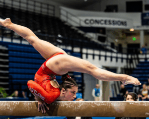 A gymnast balances on her chest holds the beam to do a backbend, with her legs in the air in a split position.