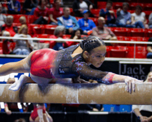 A female gymnast in a shimmering red and black long-sleeved leotard performs a balance beam routine in a packed arena. She is positioned low and horizontal to the beam, with her right arm extended forward and her left leg trailing behind. Her hair is styled in intricate braids with small silver beads, and she has a focused expression. The background shows blurred spectators in red stadium seating.