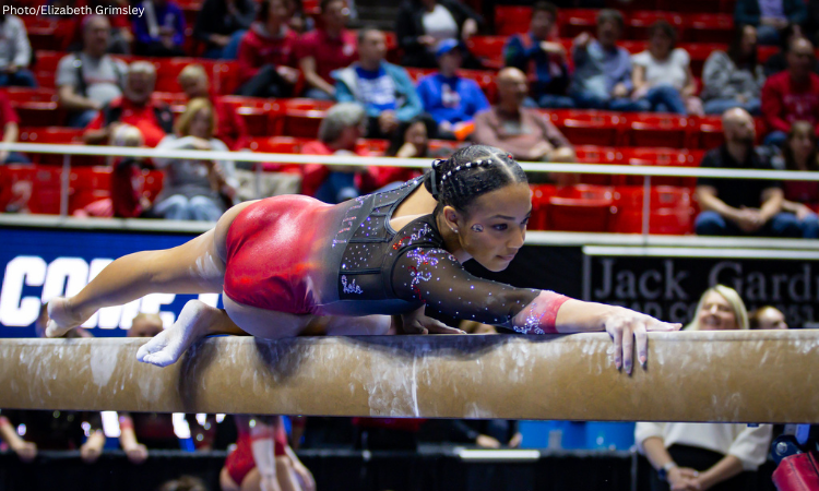 A female gymnast in a shimmering red and black long-sleeved leotard performs a balance beam routine in a packed arena. She is positioned low and horizontal to the beam, with her right arm extended forward and her left leg trailing behind. Her hair is styled in intricate braids with small silver beads, and she has a focused expression. The background shows blurred spectators in red stadium seating.