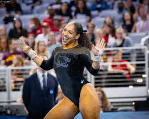 A female gymnast wearing a black long-sleeved leotard with a sparkling tiger logo on the chest and glittery accents. She has her hair pulled back in a ponytail with a black bow and is smiling broadly with her hands raised in a celebratory gesture. She is wearing white hand grips and is in a large gymnastics arena with a blurred crowd and stadium seating in the background.
