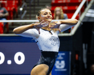 A female gymnast in a white and black bedazzled leotard performs a floor routine, holding her hands near her face with a smile. She has glitter on her temple, and the background shows blurred red stadium seating and a digital scoreboard displaying "0.0".