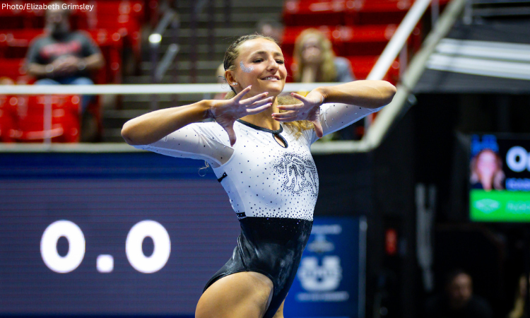 A female gymnast in a white and black bedazzled leotard performs a floor routine, holding her hands near her face with a smile. She has glitter on her temple, and the background shows blurred red stadium seating and a digital scoreboard displaying "0.0".