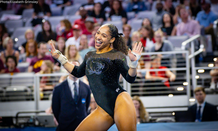 A female gymnast wearing a black long-sleeved leotard with a sparkling tiger logo on the chest and glittery accents. She has her hair pulled back in a ponytail with a black bow and is smiling broadly with her hands raised in a celebratory gesture. She is wearing white hand grips and is in a large gymnastics arena with a blurred crowd and stadium seating in the background.