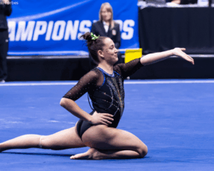 A female gymnast in a black and blue sequined leotard performs a floor routine on a blue mat. She is in a low, kneeling pose with one leg extended behind her and her left arm gracefully extended forward. The background shows a "CHAMPIONSHIPS" banner and spectators in a competition arena.
