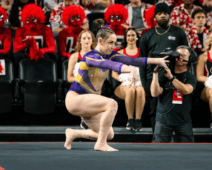 A female gymnast in a purple and gold leotard performs a floor routine during a collegiate meet. She is in a low crouch with her arms extended forward and hands posed artistically. Her ankles are wrapped in tan athletic tape. In the blurred background, several fans are dressed in full-body red paint and wigs, while a camera operator and cheerleaders watch from the sidelines.