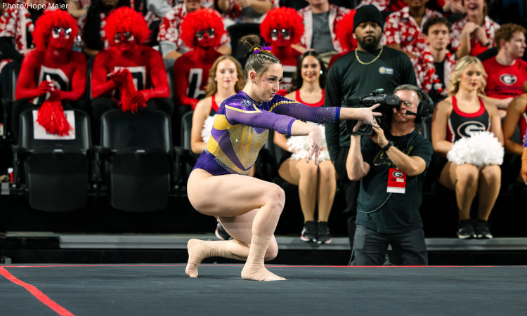 A female gymnast in a purple and gold leotard performs a floor routine during a collegiate meet. She is in a low crouch with her arms extended forward and hands posed artistically. Her ankles are wrapped in tan athletic tape. In the blurred background, several fans are dressed in full-body red paint and wigs, while a camera operator and cheerleaders watch from the sidelines.