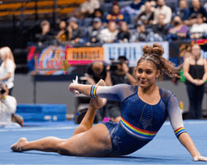 A female collegiate gymnast performing a floor routine in a crowded arena. She is lying on the blue floor mat in a graceful, arched pose, looking toward the camera with a smile. She wears a navy blue long-sleeved leotard featuring sparkling rhinestones and vibrant rainbow-striped bands on the chest and sleeves. Her hair is styled in a high curly bun with braided details. The background shows a blurred audience in the stands and event staff.