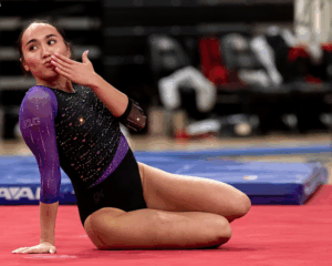 A female collegiate gymnast is sitting on a red floor mat in a posing position at the conclusion of a floor routine. She is leaning back on one hand while blowing a kiss toward the side. She is wearing a black and purple long-sleeved leotard embellished with shimmering rhinestones and a "B1G" logo on the sleeve. The background shows a blurred gymnasium setting with blue landing mats and empty chairs.