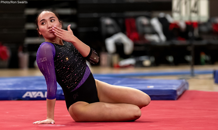 A female collegiate gymnast is sitting on a red floor mat in a posing position at the conclusion of a floor routine. She is leaning back on one hand while blowing a kiss toward the side. She is wearing a black and purple long-sleeved leotard embellished with shimmering rhinestones and a "B1G" logo on the sleeve. The background shows a blurred gymnasium setting with blue landing mats and empty chairs.