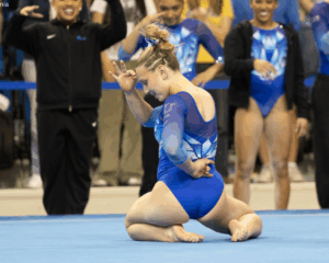 A gymnast in a blue and white long-sleeved leotard performs a floor routine on a blue mat. She is kneeling with one arm tucked behind her back and the other raised to her forehead, fingers in an "OK" gesture. In the background, teammates and coaches in matching blue athletic gear watch and cheer.
