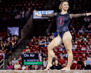 An image of a Stanford gymnast, Anna Roberts, performing on the balance beam in a crowded arena. She is captured in a graceful pose, standing on her right leg with her left heel slightly lifted, her arms extended out to her sides, and a focused smile on her face. She is wearing a black long-sleeved leotard featuring a large, sparkling red and white Stanford "S" logo on the chest. In the background, a digital "On Air" scoreboard identifies her as "A. ROBERTS" on the "BALANCE BEAM," and the stadium stands are filled with spectators.