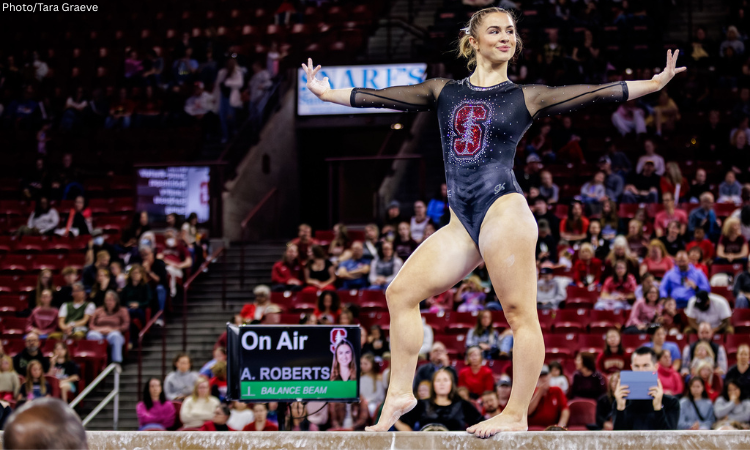 An image of a Stanford gymnast, Anna Roberts, performing on the balance beam in a crowded arena. She is captured in a graceful pose, standing on her right leg with her left heel slightly lifted, her arms extended out to her sides, and a focused smile on her face. She is wearing a black long-sleeved leotard featuring a large, sparkling red and white Stanford "S" logo on the chest. In the background, a digital "On Air" scoreboard identifies her as "A. ROBERTS" on the "BALANCE BEAM," and the stadium stands are filled with spectators.