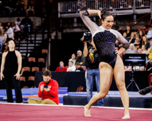 An action shot of a female gymnast performing a floor routine in a crowded arena. She is positioned in the center of the frame, striking a pose with one hand behind her head and the other on her hip. She wears a black leotard with sheer, long sleeves and shimmering purple and silver embellishments across the chest. In the blurred background, spectators fill the tiered seating of the stadium. Several photographers and staff members are visible near the edge of the maroon gymnastics floor, including a man in a red sweatshirt kneeling to take a photo and a woman in a black sleeveless top standing nearby. A small watermark in the top-left corner reads "Photo/Tara Graeve."