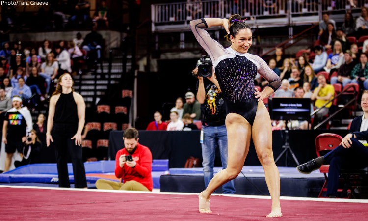 An action shot of a female gymnast performing a floor routine in a crowded arena. She is positioned in the center of the frame, striking a pose with one hand behind her head and the other on her hip. She wears a black leotard with sheer, long sleeves and shimmering purple and silver embellishments across the chest. In the blurred background, spectators fill the tiered seating of the stadium. Several photographers and staff members are visible near the edge of the maroon gymnastics floor, including a man in a red sweatshirt kneeling to take a photo and a woman in a black sleeveless top standing nearby. A small watermark in the top-left corner reads "Photo/Tara Graeve."