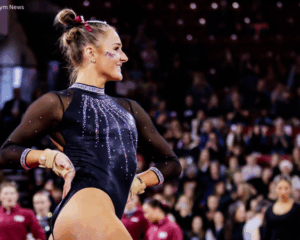 Sophia Isbell, a gymnast from Texas Woman's University (TWU) stands in profile, smiling toward the right in a brightly lit arena. She is wearing a black long-sleeved leotard embellished with shimmering silver rhinestones in a waterfall pattern across the chest and around the neck and cuffs. Her hair is styled in a high bun with a maroon bow, and she has a "TWU" temporary tattoo on her cheek. She wears tan gymnastics grips on her hands, which are resting on her hips as she poses. The background is a blurred audience in a stadium setting.