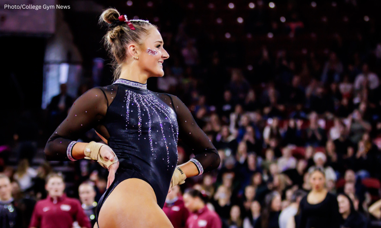 Sophia Isbell, a gymnast from Texas Woman's University (TWU) stands in profile, smiling toward the right in a brightly lit arena. She is wearing a black long-sleeved leotard embellished with shimmering silver rhinestones in a waterfall pattern across the chest and around the neck and cuffs. Her hair is styled in a high bun with a maroon bow, and she has a "TWU" temporary tattoo on her cheek. She wears tan gymnastics grips on her hands, which are resting on her hips as she poses. The background is a blurred audience in a stadium setting.