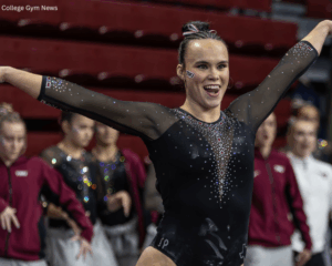 Sophie Hernandez, a female collegiate gymnast for Texas Woman's University (TWU), smiles brightly with her arms outstretched in a floor routine pose. She is wearing a black, long-sleeved leotard embellished with silver and red crystals, with a TWU logo temporary tattoo on her cheek. In the blurred background, her teammates in maroon warm-up jackets and black leotards cheer her on from the sidelines of the arena.
