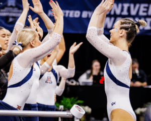 Arizona gymnasts, including Alysen Fears, celebrate after a routine, raising their arms for high-fives while wearing white and navy leotards.