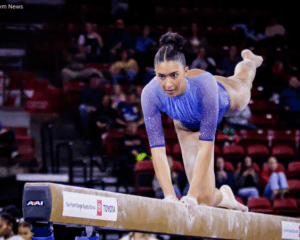 An action shot of a gymnast, Zoe Claiborne from LIU, performing on a balance beam. She is wearing a shimmering light blue leotard with sheer sleeves. She is positioned on her right knee on the beam, leaning forward with her hands gripping the edges and her left leg extended straight back. Her expression is focused as she looks ahead. The background shows a blurred audience in a stadium with red seating. The beam features a Toyota logo and an AAI equipment mark.
