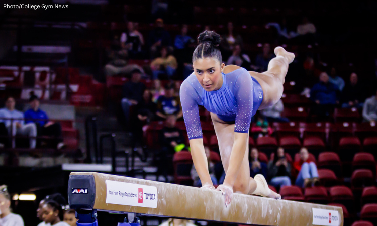 An action shot of a gymnast, Zoe Claiborne from LIU, performing on a balance beam. She is wearing a shimmering light blue leotard with sheer sleeves. She is positioned on her right knee on the beam, leaning forward with her hands gripping the edges and her left leg extended straight back. Her expression is focused as she looks ahead. The background shows a blurred audience in a stadium with red seating. The beam features a Toyota logo and an AAI equipment mark.