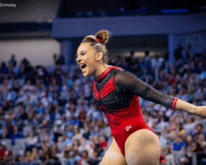 Makenna Smith celebrates with arms outstretched and a wide smile on the floor, wearing a red Utah leotard as the crowd fills the arena behind her.