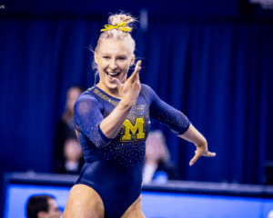 A blonde female gymnast wearing a dark blue and gold Michigan leotard is smiling and striking a dynamic pose on the floor exercise. Her hair is pulled up in a bun, and she has an M sticker on her cheekbones. The background is a blurred navy blue, suggesting an indoor arena.