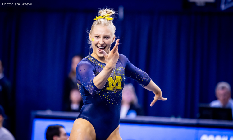 A blonde female gymnast wearing a dark blue and gold Michigan leotard is smiling and striking a dynamic pose on the floor exercise. Her hair is pulled up in a bun, and she has an M sticker on her cheekbones. The background is a blurred navy blue, suggesting an indoor arena.