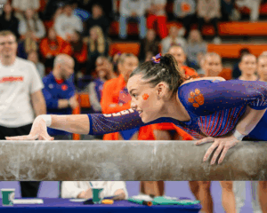 Emma Malewski leans low over the balance beam with one hand extended forward, focused and smiling slightly. She wears a purple Clemson leotard with orange paw-print accents as teammates and coaches watch from the background.