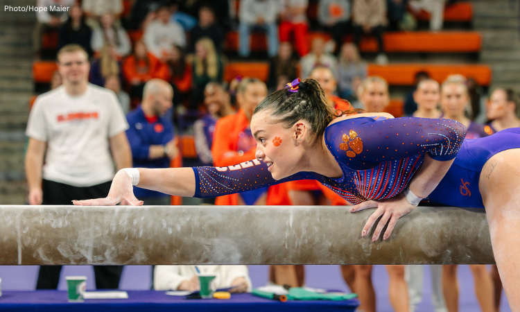 Emma Malewski leans low over the balance beam with one hand extended forward, focused and smiling slightly. She wears a purple Clemson leotard with orange paw-print accents as teammates and coaches watch from the background.