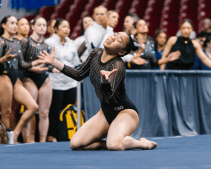 Aurélie Tran performs a floor routine, kneeling on the mat with her arms extended and eyes closed in an expressive moment. She wears a black leotard with sparkling vertical rhinestone lines as teammates and coaches watch from the sideline in the background.