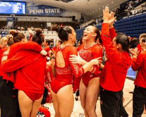 A group of female gymnasts, wearing red leotards and warm-up gear, hug and celebrate excitedly in a large indoor arena after a competition.