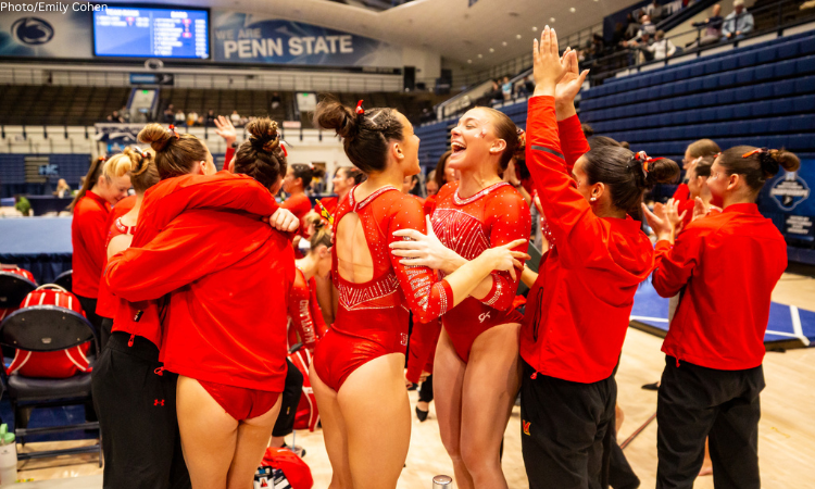 A group of female gymnasts, wearing red leotards and warm-up gear, hug and celebrate excitedly in a large indoor arena after a competition.