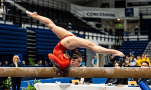 Maryland gymnast Maddie Komoroski shows off her flexibility on beam with a chest stand and a split. She is wearing a red leotard with "Maryland" on the sleeve.