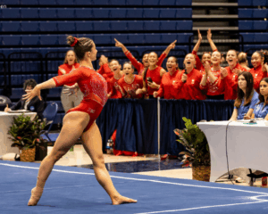 A gymnast, wearing all red, poses in a lunge and with her arms back in the corner of the floor exercise. Her team cheers from the sidelines.