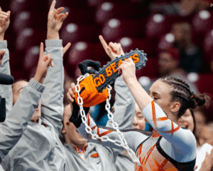 A gymnast celebrates with her arms up and a team around her, lifting a crocheted chainsaw that says "Go Beavs". She wears an orange leotard with white and orange striped sleeves, and her hair is pulled back with braids. Her eyes are closed.
