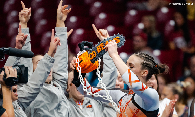 A gymnast celebrates with her arms up and a team around her, lifting a crocheted chainsaw that says "Go Beavs". She wears an orange leotard with white and orange striped sleeves, and her hair is pulled back with braids. Her eyes are closed.