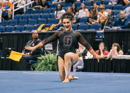 Sienna Robinson poses during her floor routine. She is smiling and on one knee with her arms to the side. She is wearing a black leotard with a red Stanford "S" on the chest.