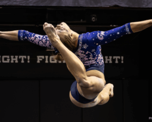 A gymnast mid-leap, viewed from behind, with her leg pointed back to her head in a ring position and arms out to the side. She is in front of a black background and wears a blue leotard with pink flowers.