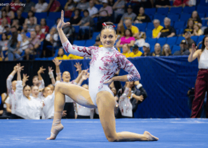 Gabby Gladieux of Alabama performs a floor routine, posed on one knee with one arm raised and the other on her hip, wearing a white leotard with red detailing. Teammates cheer in the background as a crowd watches from blue arena seating.