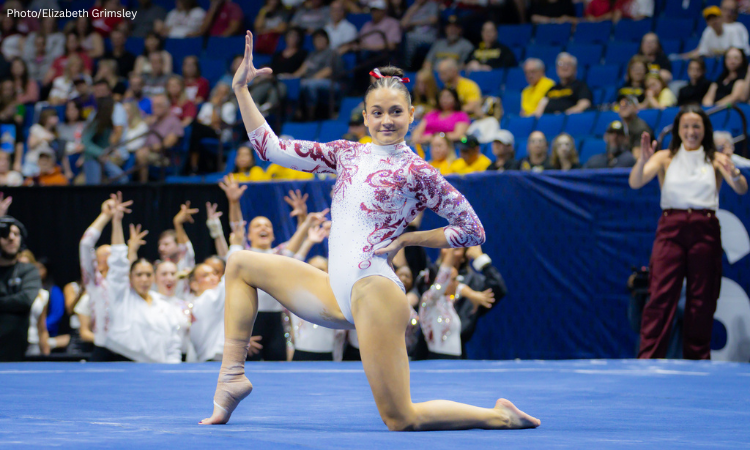Gabby Gladieux of Alabama performs a floor routine, posed on one knee with one arm raised and the other on her hip, wearing a white leotard with red detailing. Teammates cheer in the background as a crowd watches from blue arena seating.