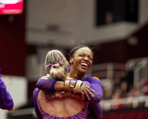 Clemson gymnast Brie Clark embraces a teammate in a close hug during competition, both wearing purple leotards. Clark is facing the camera with a wide, joyful smile and eyes closed in celebration, while her teammate’s back is turned toward the camera. The background shows a blurred arena setting with spectators and team staff on the sidelines.